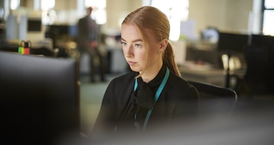 Woman dressed in a black blouse, in an office seated working in front of a computer screen.
